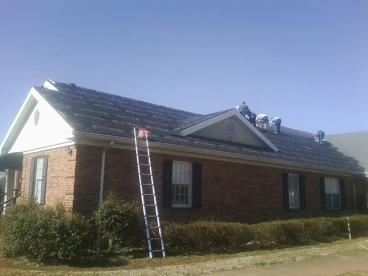 Crew tearing shingles off a brick ranch roof with a ladder set — Metal Roof Repair work by our crew in Clyde